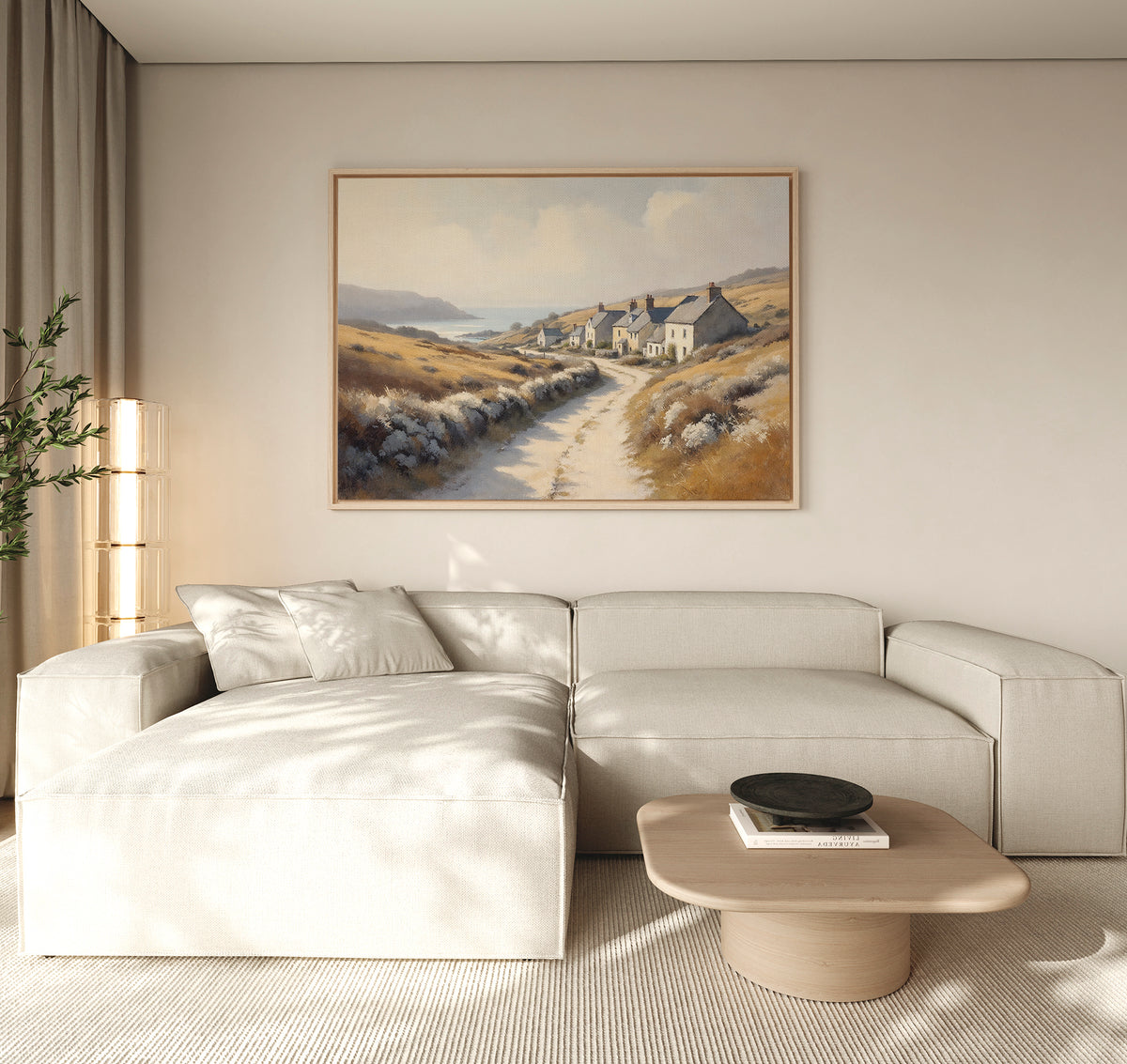 A modern living room features a light beige sectional, a low wooden coffee table with a black bowl, and the Coastal Haven Canvas Art - Horizontal depicting a country path by the sea. A tall plant sits in the corner near floor-length curtains.