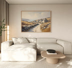A modern living room features a light beige sectional, a low wooden coffee table with a black bowl, and the Coastal Haven Canvas Art - Horizontal depicting a country path by the sea. A tall plant sits in the corner near floor-length curtains.