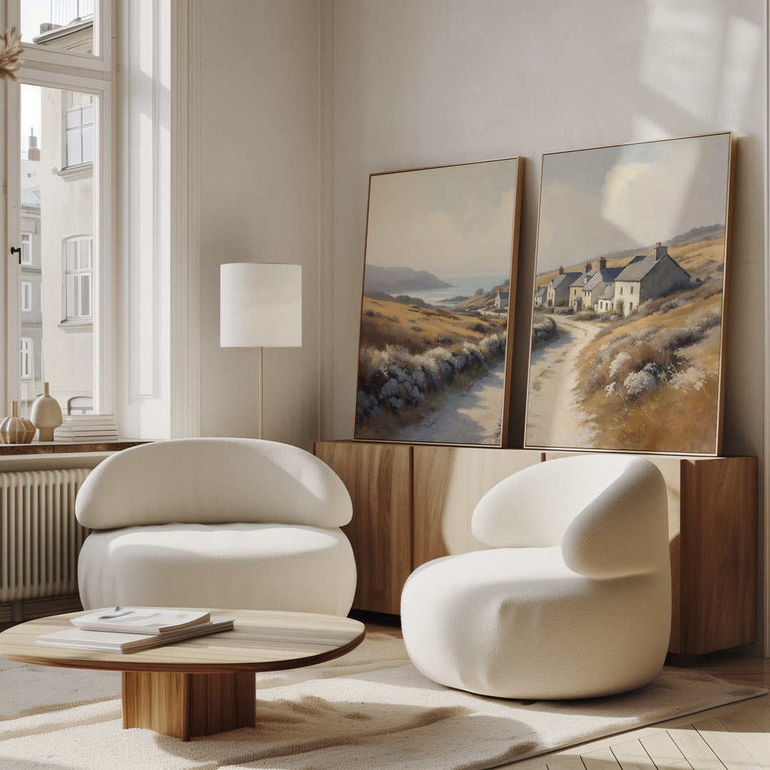 Modern living room with two curved white chairs, a low wooden table, and the Coastal Haven - Set of 2 landscape canvas prints on a wooden credenza. Large windows bring natural light into this cozy, rustic-inspired minimalist space.