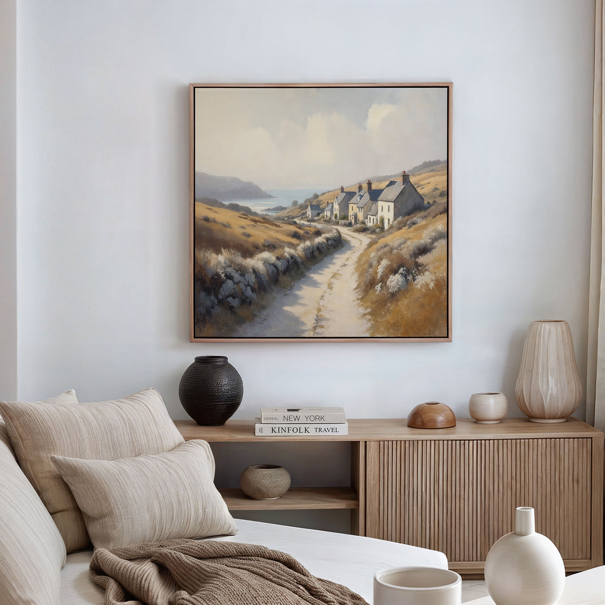 A cozy living room with a light sofa, neutral cushions, a wooden sideboard with vases and books, and the Coastal Haven Canvas Art - Square, showcasing a rustic coastal landscape above.