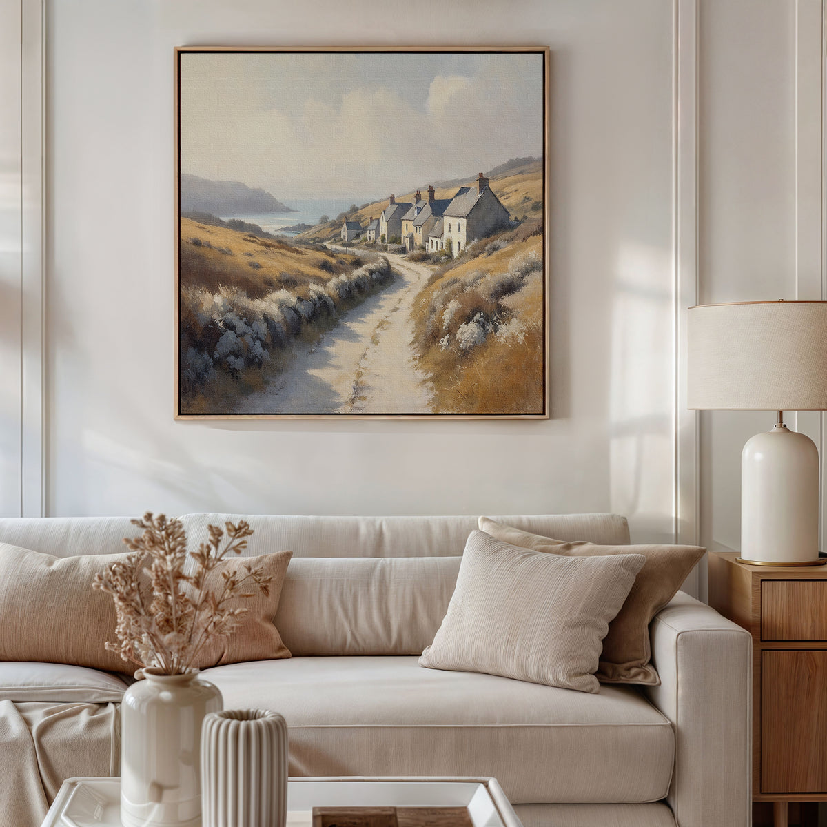 A cozy living room with a white sofa, beige cushions, a side table with a lamp, vases, and dried flowers. Above the sofa hangs the Coastal Haven Canvas Art - Square, featuring a coastal landscape with cottages and a winding path.