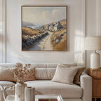 A cozy living room with a white sofa, beige cushions, a side table with a lamp, vases, and dried flowers. Above the sofa hangs the Coastal Haven Canvas Art - Square, featuring a coastal landscape with cottages and a winding path.