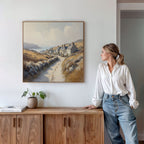 A woman in a white blouse and jeans leans against a wooden cabinet, gazing at the Coastal Haven Canvas Art - Square displayed on the white wall above her. A potted plant and books are arranged on top of the cabinet.