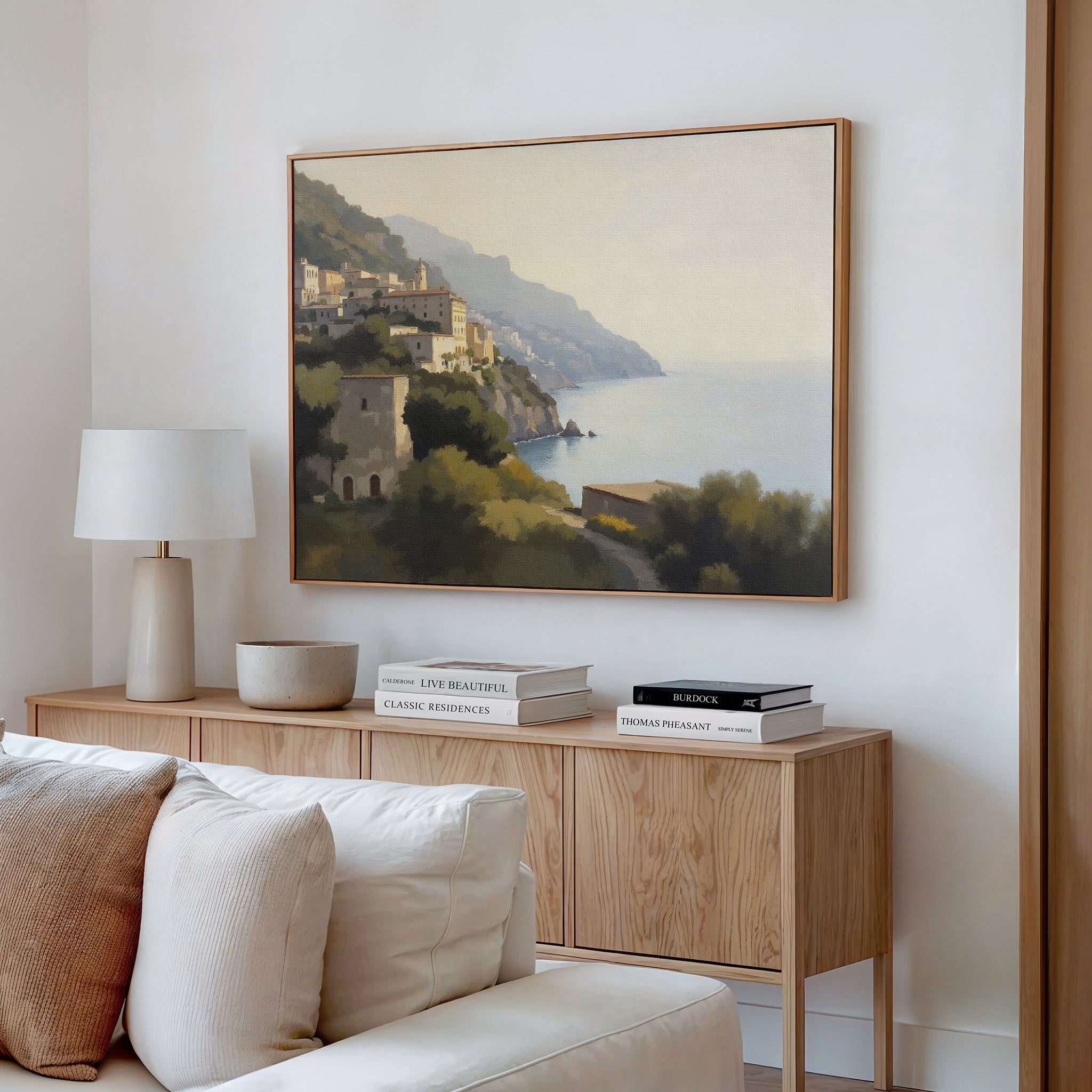 A minimal living room with a light wood console, white lamp, neutral decor, and the Italian Reverie Canvas Art - Horizontal framed above the console. Books and ceramic pieces are neatly arranged on the console.
