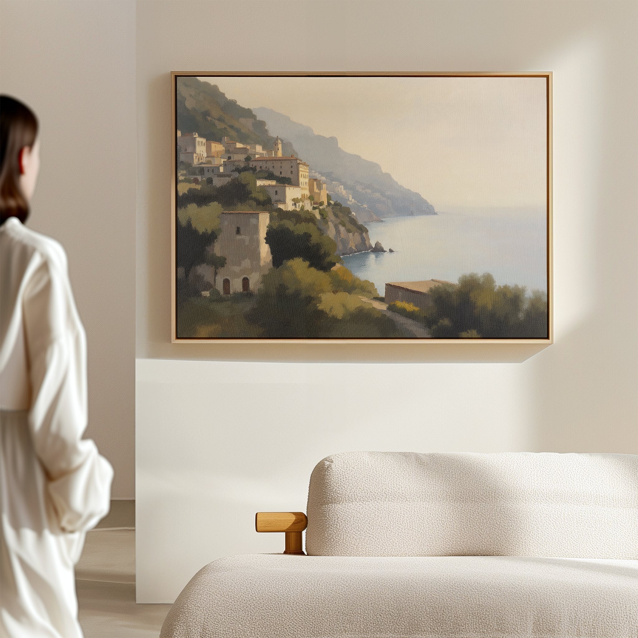 A woman in a white robe stands in a minimalist room, admiring the Italian Reverie Canvas Art - Horizontal depicting Mediterranean cliffside architecture and sea cliffs above a light-colored sofa.
