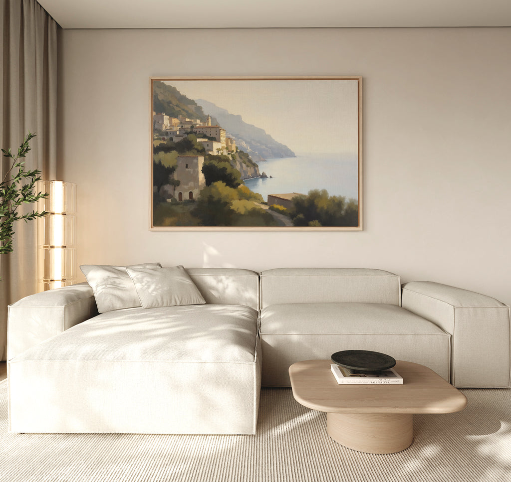A modern living room with a light beige sectional sofa, round wooden coffee table, potted plant, and the Italian Reverie Canvas Art - Horizontal depicting a coastal village. Soft neutrals create a calm, inviting atmosphere.