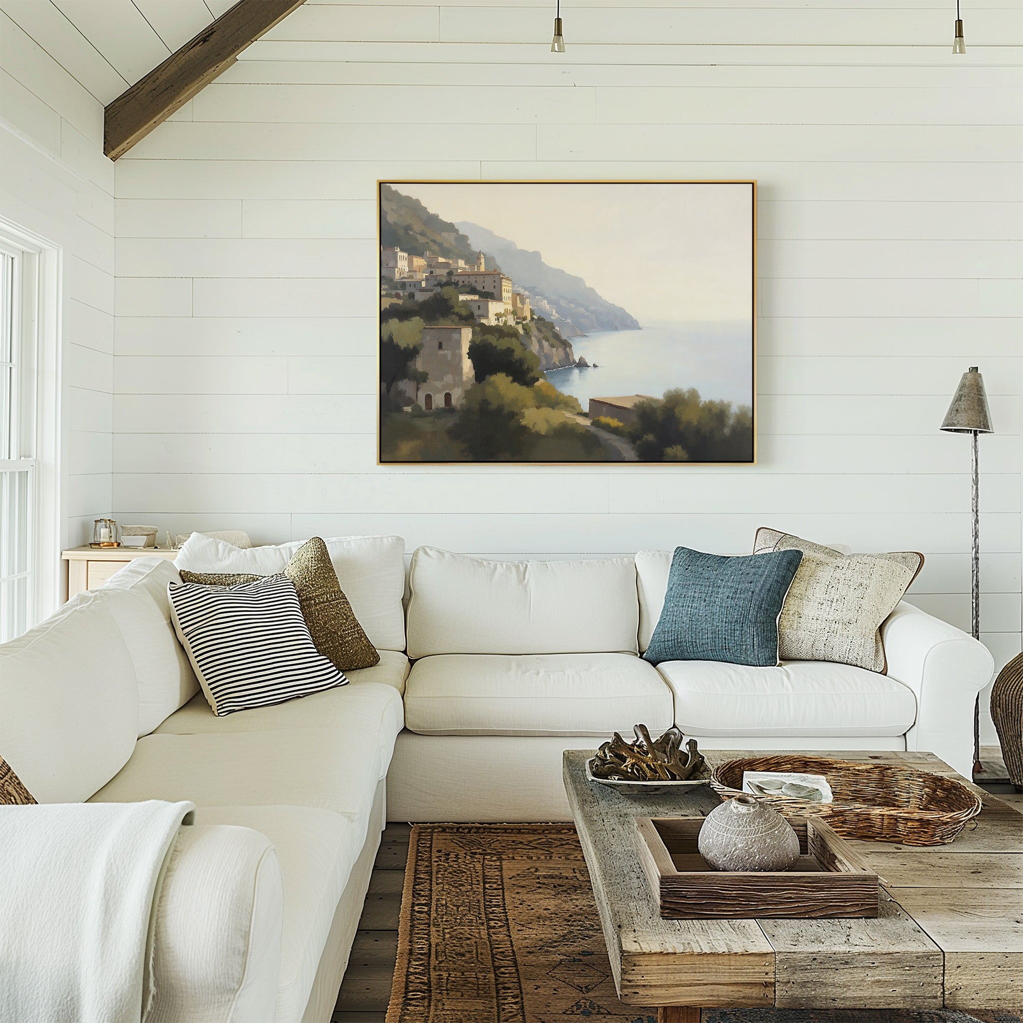 A cozy living room features a white sectional sofa, assorted pillows, a rustic wooden coffee table, and the Italian Reverie Canvas Art - Horizontal depicting Mediterranean cliffside views. Natural light pours in from tall windows.