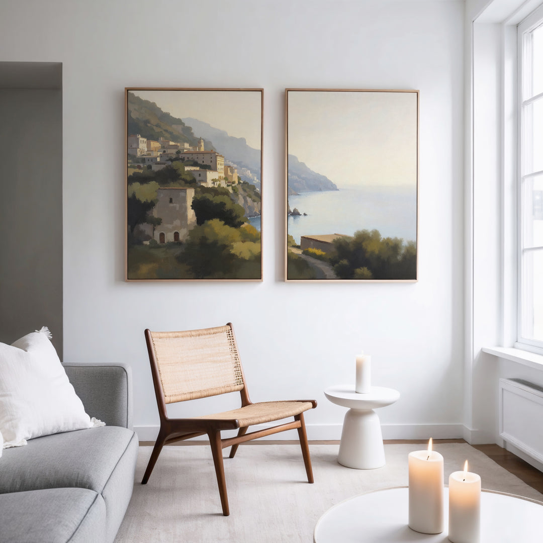 A modern living room with a light gray sofa, woven chair, and round white table with a candle. Two large "Italian Reverie - Set of 2" coastal wall art pieces add elegance. Three candles glow on the small side table.