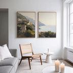 A modern living room with a light gray sofa, woven chair, and round white table with a candle. Two large "Italian Reverie - Set of 2" coastal wall art pieces add elegance. Three candles glow on the small side table.