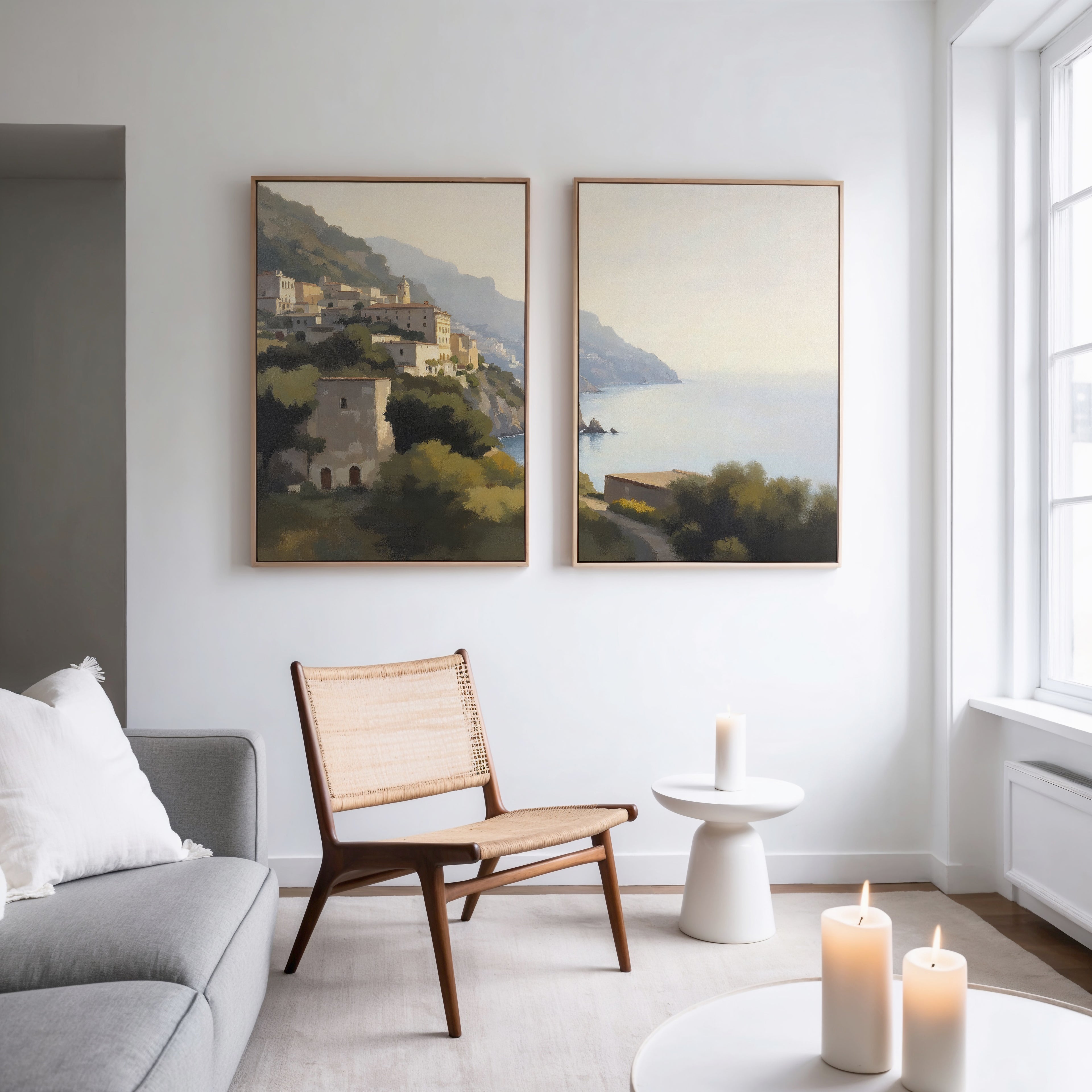A modern living room with a light gray sofa, woven chair, and round white table with a candle. Two large "Italian Reverie - Set of 2" coastal wall art pieces add elegance. Three candles glow on the small side table.