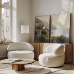 A bright, modern living room features two curvy white armchairs, a round wooden coffee table, a wooden sideboard, and the "Italian Reverie - Set of 2" landscape paintings leaning beneath tall windows for elegant decor.