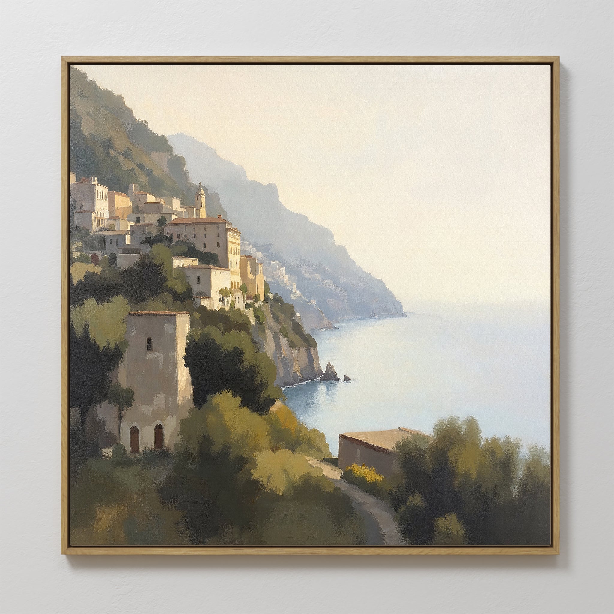 Italian Reverie Canvas Art - Square: A framed Mediterranean print depicting a coastal hillside village with sunlit buildings, lush greenery, and calm blue sea under a clear sky—the perfect touch of elegance for your home decor.