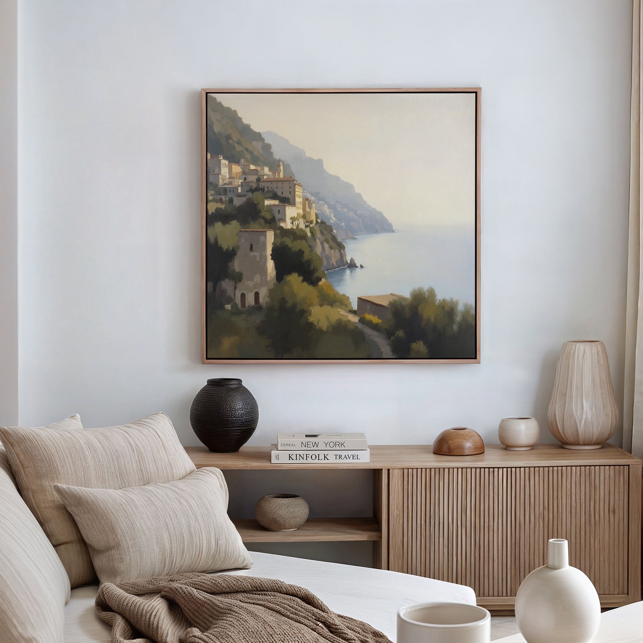 A cozy, modern living room features a neutral-toned sofa, a wooden sideboard with elegant decor, and the Italian Reverie Canvas Art - Square depicting a Mediterranean coastal town hanging on the white wall.