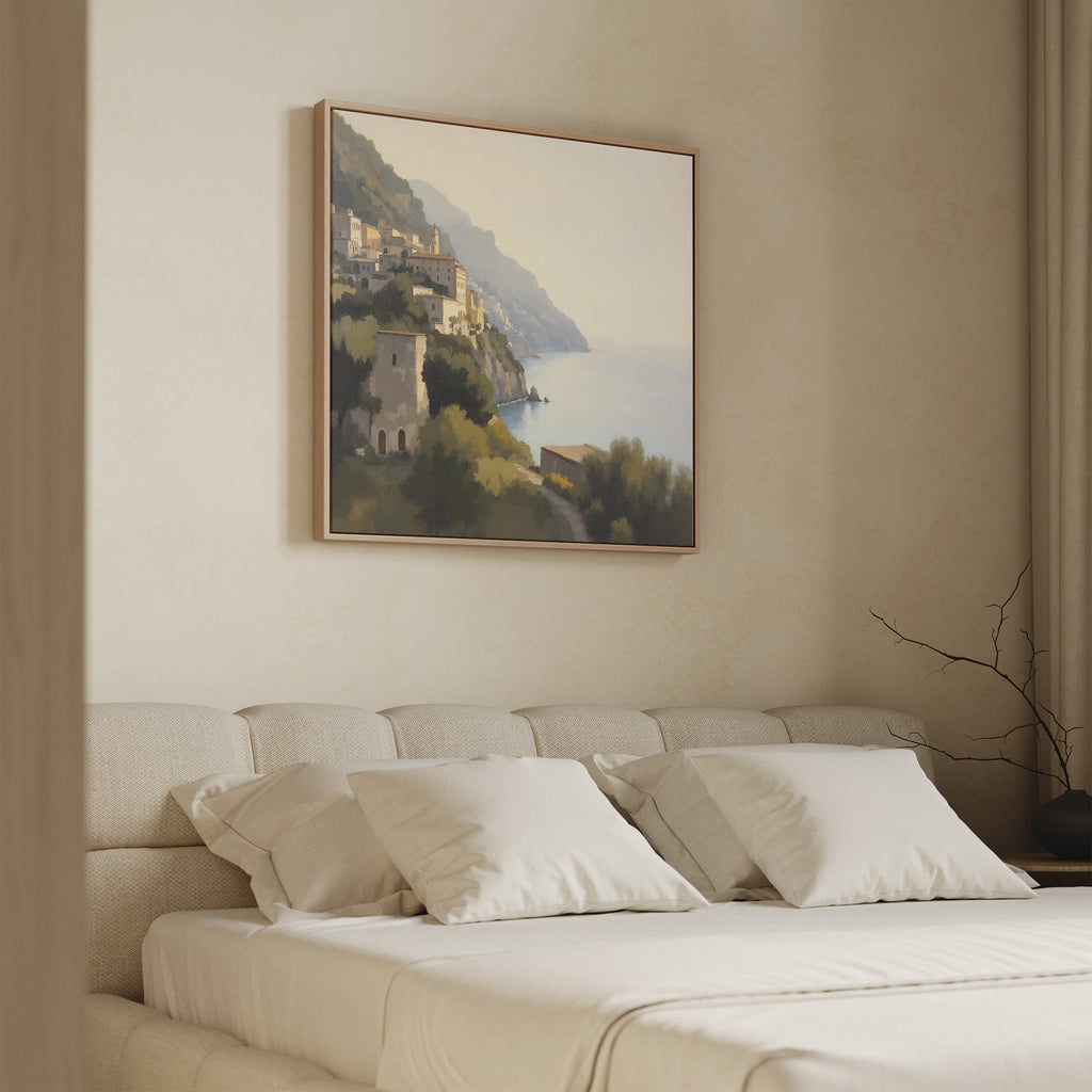 A beige, minimalist bedroom features a low upholstered bed with white pillows. Above, the Italian Reverie Canvas Art - Square depicts a Mediterranean village under a hazy sky, adding elegance. A bare branch graces the nightstand.
