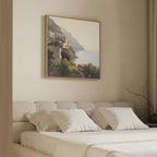 A beige, minimalist bedroom features a low upholstered bed with white pillows. Above, the Italian Reverie Canvas Art - Square depicts a Mediterranean village under a hazy sky, adding elegance. A bare branch graces the nightstand.