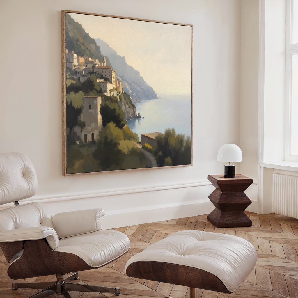 A modern living room featuring a white lounge chair and ottoman, a wooden side table with a white lamp, and the Italian Reverie Canvas Art - Square—a large landscape of a Mediterranean town—displayed near a spacious window.
