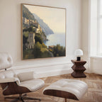 A modern living room featuring a white lounge chair and ottoman, a wooden side table with a white lamp, and the Italian Reverie Canvas Art - Square—a large landscape of a Mediterranean town—displayed near a spacious window.