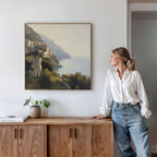 A woman in a white blouse and jeans leans on a wooden sideboard, admiring the Italian Reverie Canvas Art - Square. A potted plant and books enhance the tranquil, white-walled room’s cozy, elegant vibe.