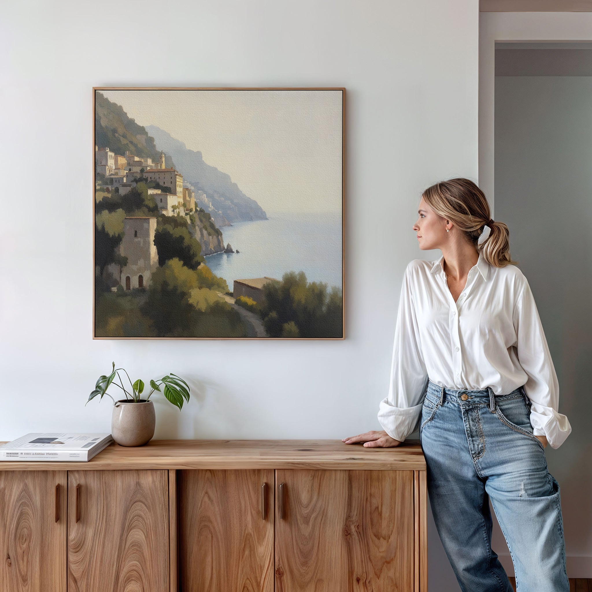 A woman in a white blouse and jeans leans on a wooden sideboard, admiring the Italian Reverie Canvas Art - Square. A potted plant and books enhance the tranquil, white-walled room’s cozy, elegant vibe.