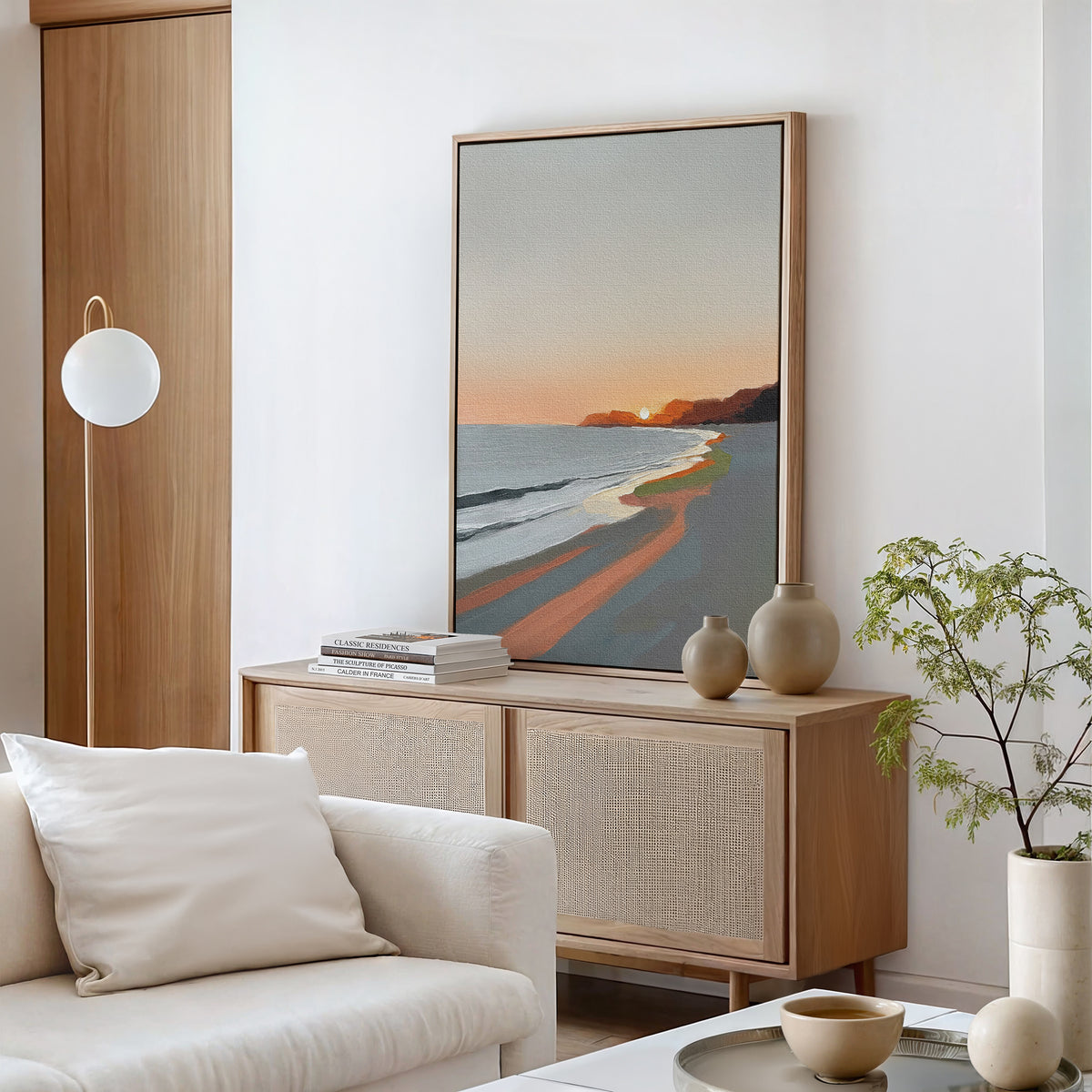 A modern living room with neutral tones features Coastal Reverie Canvas Art - Vertical, a light wood sideboard with vases and books, a white sofa, and a small green plant for tranquil home decor.