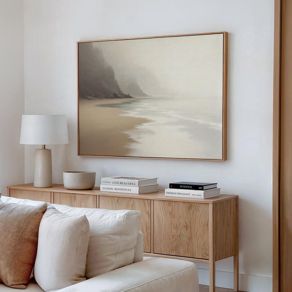 A modern living room with the Coastal Silence Canvas Art - Horizontal depicting a foggy beach above a cream sofa, light wood sideboard, stacked books, lamp, and bowl—ideal for those who love muted coastal decor.