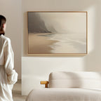 A person in white stands in a minimalist room, gazing at the Coastal Silence Canvas Art - Horizontal featuring a misty beach with waves and cliffs, above a light-colored sofa, evoking muted coastal decor.