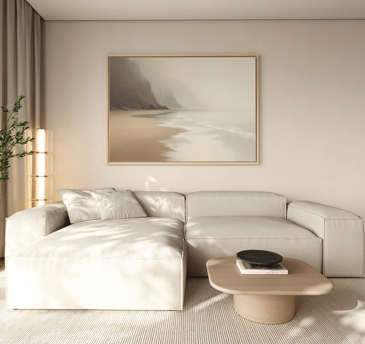 A minimalist living room features a light beige sectional, low wood coffee table, and the Coastal Silence Canvas Art - Horizontal depicting a misty beach. Muted coastal decor and soft lighting create a calm, serene ambiance.
