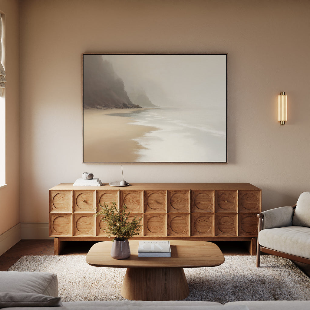 A cozy living room with neutral tones features a wooden sideboard, a simple coffee table with flowers, beige sofas on a light rug, and the Coastal Silence Canvas Art - Horizontal as an abstract coastal focal point. Soft lighting adds to the calm atmosphere.
