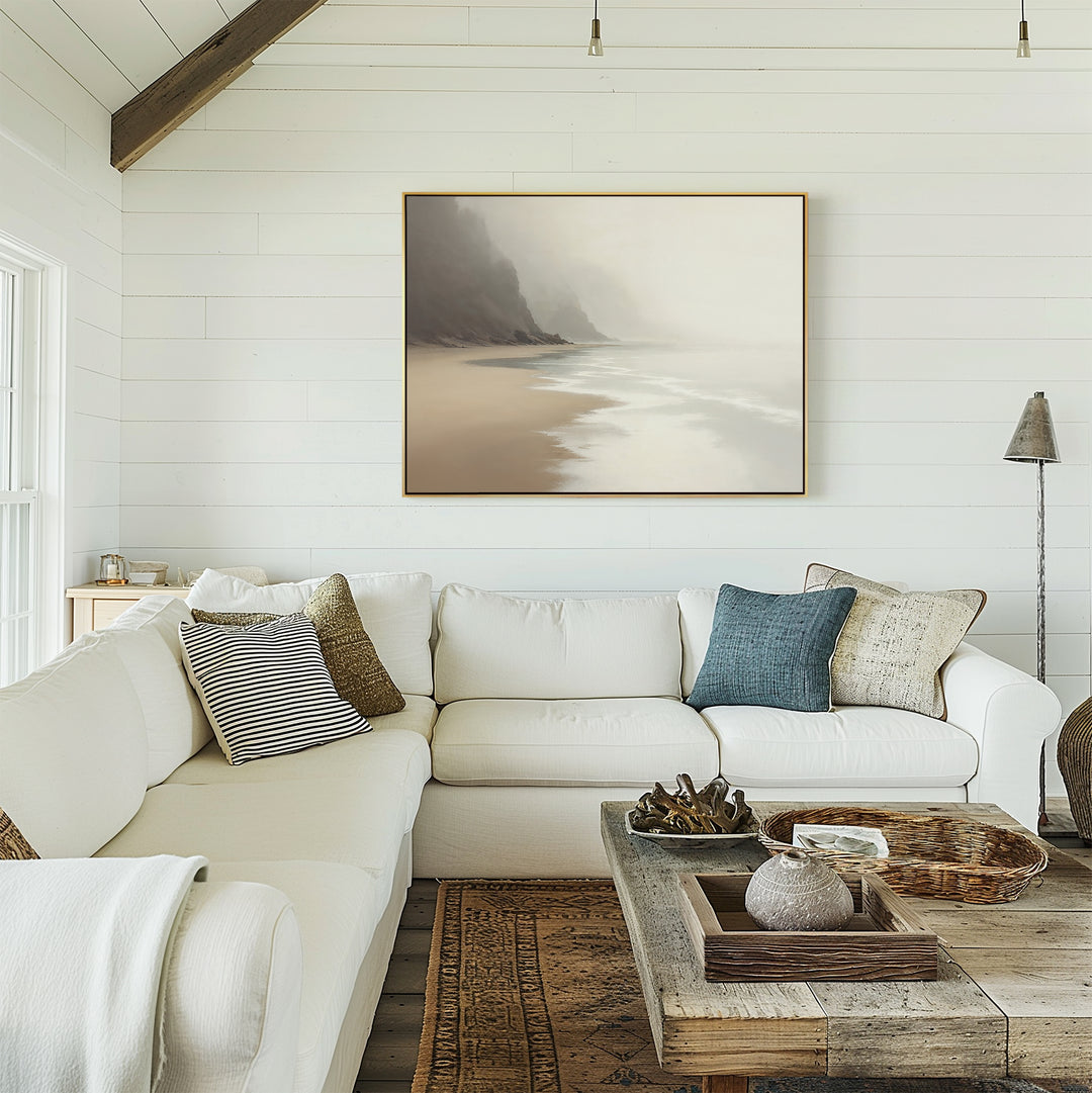 A cozy living room features a white sectional, neutral pillows, rustic wood coffee table, and muted coastal accents. The "Coastal Silence Canvas Art - Horizontal" depicting a misty beach scene hangs on a white shiplap wall.