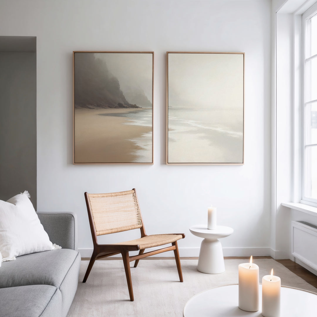 A bright, minimalist living room features a gray sofa, wooden chair, and the "Coastal Silence - Set of 2" wall art above. A small white side table with a candle and more candles on a round table by the window complete the serene Nordic Park vibe.
