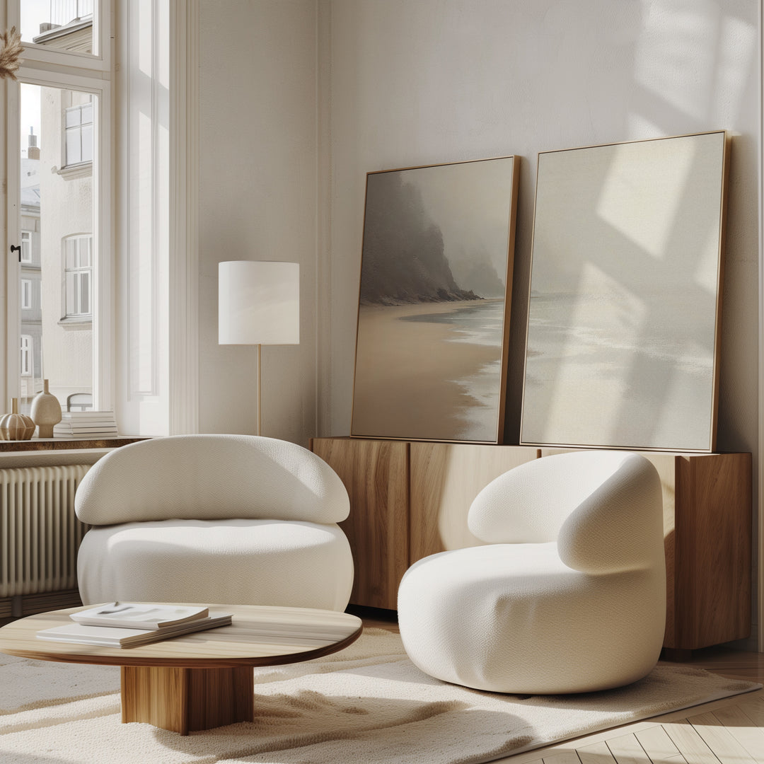 A bright, minimalist living room with two modern white armchairs, a low wooden coffee table, a wooden sideboard, and the "Coastal Silence - Set of 2" canvas prints leaning against the wall. Sunlight streams through tall windows.