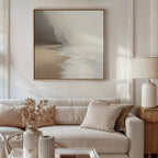 A neutral-toned living room features a beige sofa with cream pillows, a wooden side table with a white lamp, dried flowers in a vase, and the Coastal Silence Canvas Art - Square above the sofa for a serene, natural palette.
