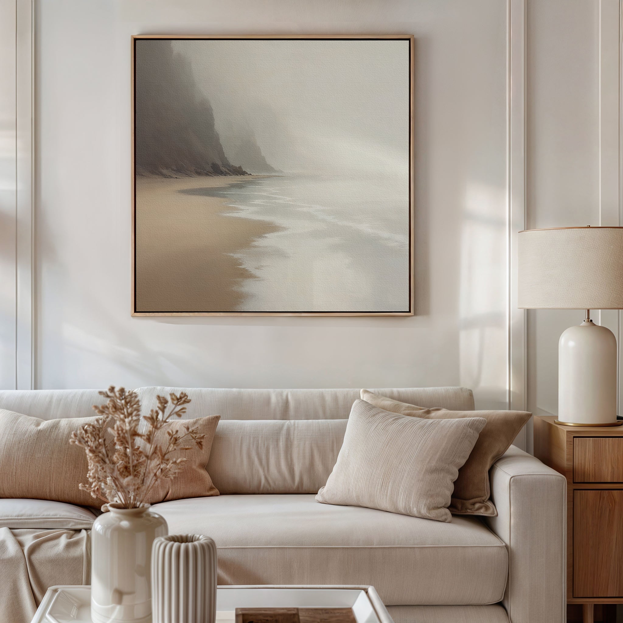 A neutral-toned living room features a beige sofa with cream pillows, a wooden side table with a white lamp, dried flowers in a vase, and the Coastal Silence Canvas Art - Square above the sofa for a serene, natural palette.
