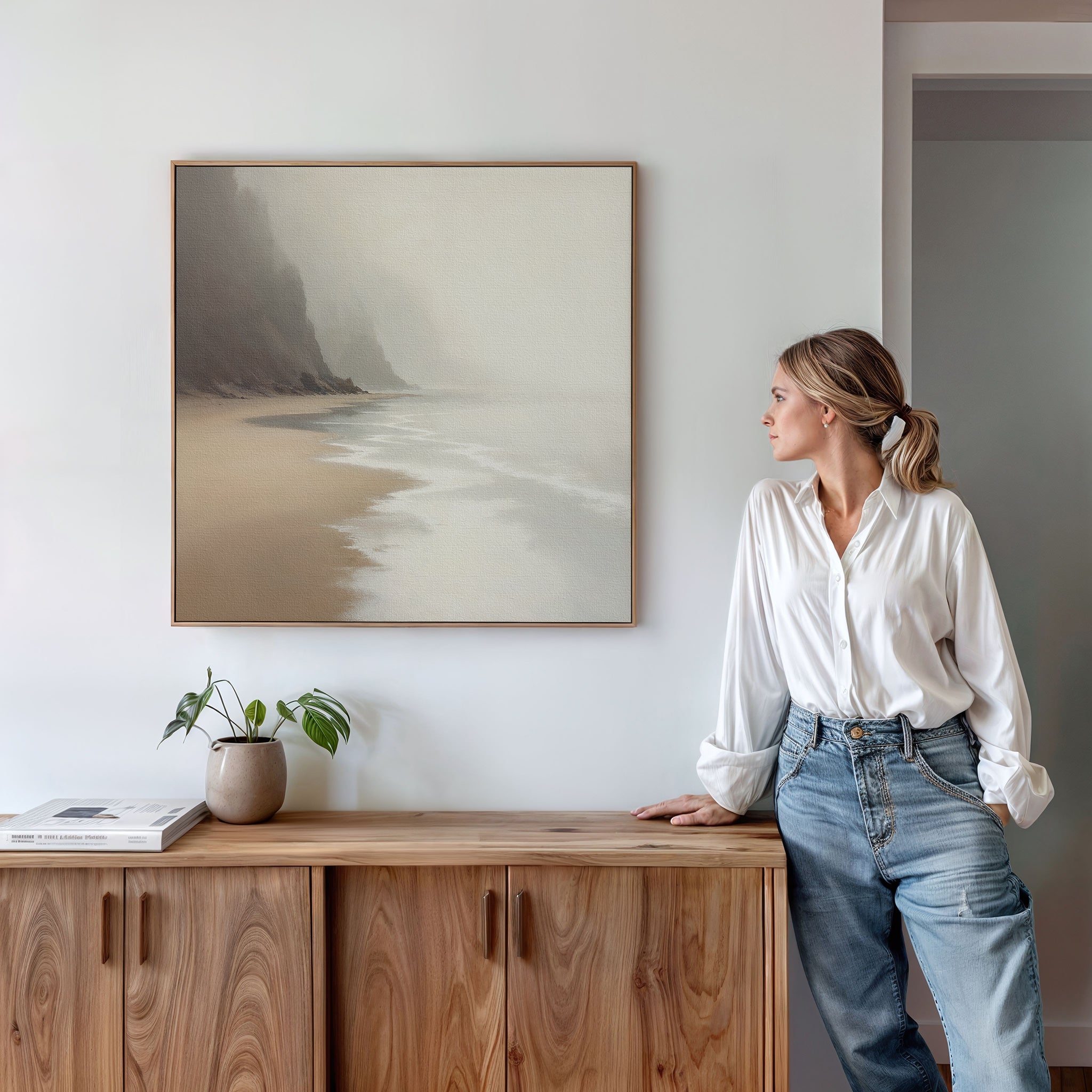 A woman in a white shirt and jeans stands by a wooden cabinet, admiring the Coastal Silence Canvas Art - Square. A small potted plant and books on the cabinet enhance the room’s natural palette and serene feel.