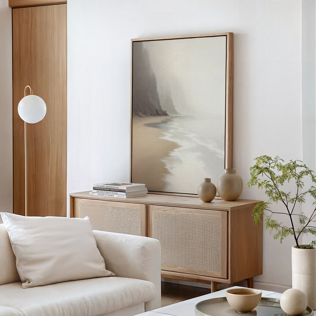 A minimalist living room featuring light wood furniture, neutral ceramics, a stack of books, a leafy plant, a modern round wall lamp, and the Coastal Silence Canvas Art - Vertical—all in soft, natural colors.