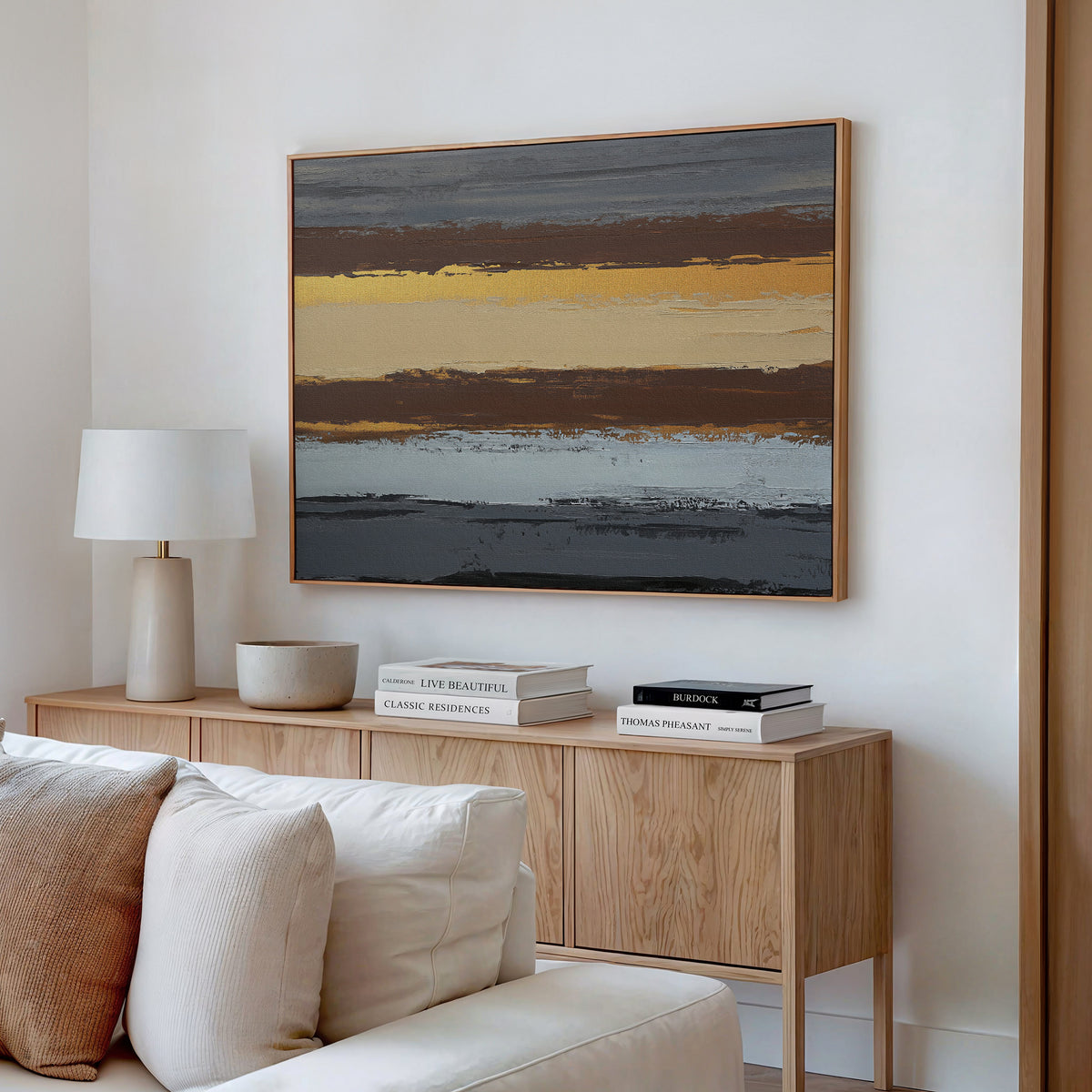A modern living room features a wooden sideboard with books, a lamp, and bowl, topped by the Coastal Strata Canvas Art - Horizontal in yellow, gray, and brown. A cream sofa is partially visible, adding earthy sophistication to the space.