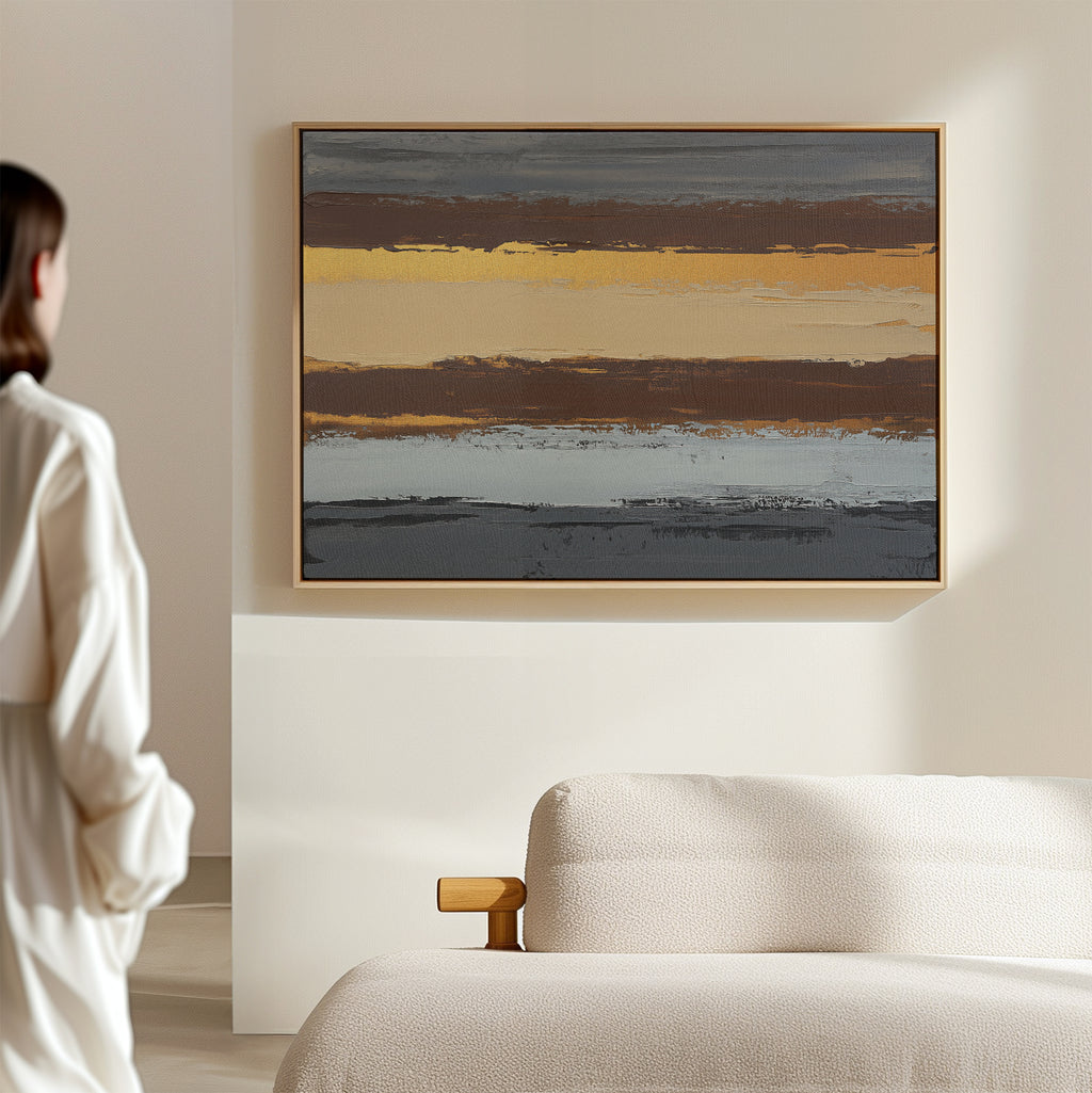 A person in a white shirt stands in a bright, minimal room facing the Coastal Strata Canvas Art - Horizontal, an abstract painting with earthy horizontal stripes, displayed above a light-colored textured sofa.