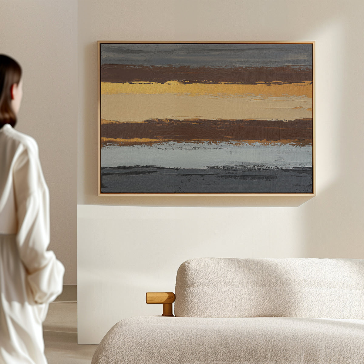 A person in a white shirt stands in a bright, minimal room facing the Coastal Strata Canvas Art - Horizontal, an abstract painting with earthy horizontal stripes, displayed above a light-colored textured sofa.