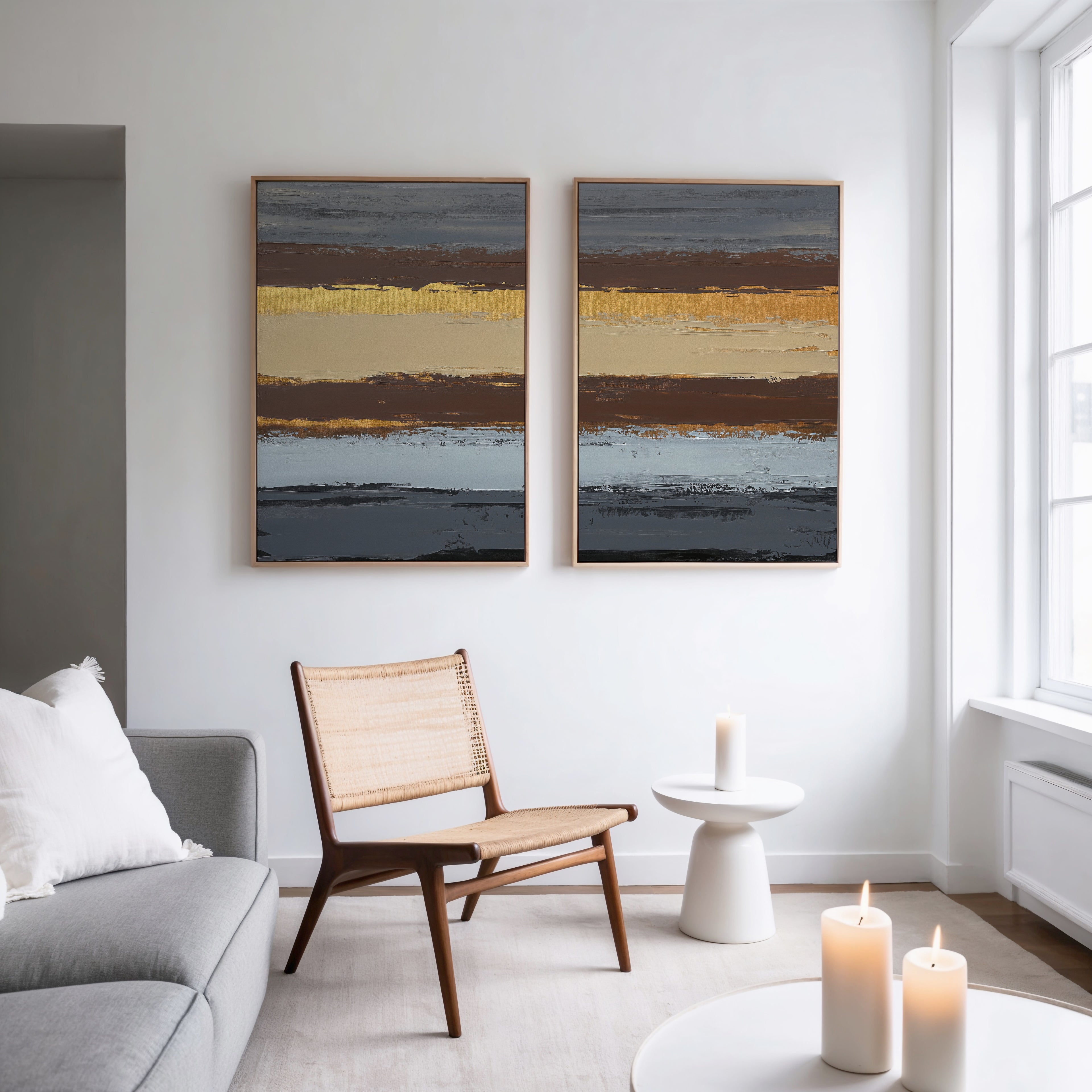 A modern living room features a gray sofa, a woven wooden chair, and Coastal Strata - Set Of 2 abstract paintings with horizontal stripes. A round side table with a candle and large windows enhance the room’s earthy sophistication.