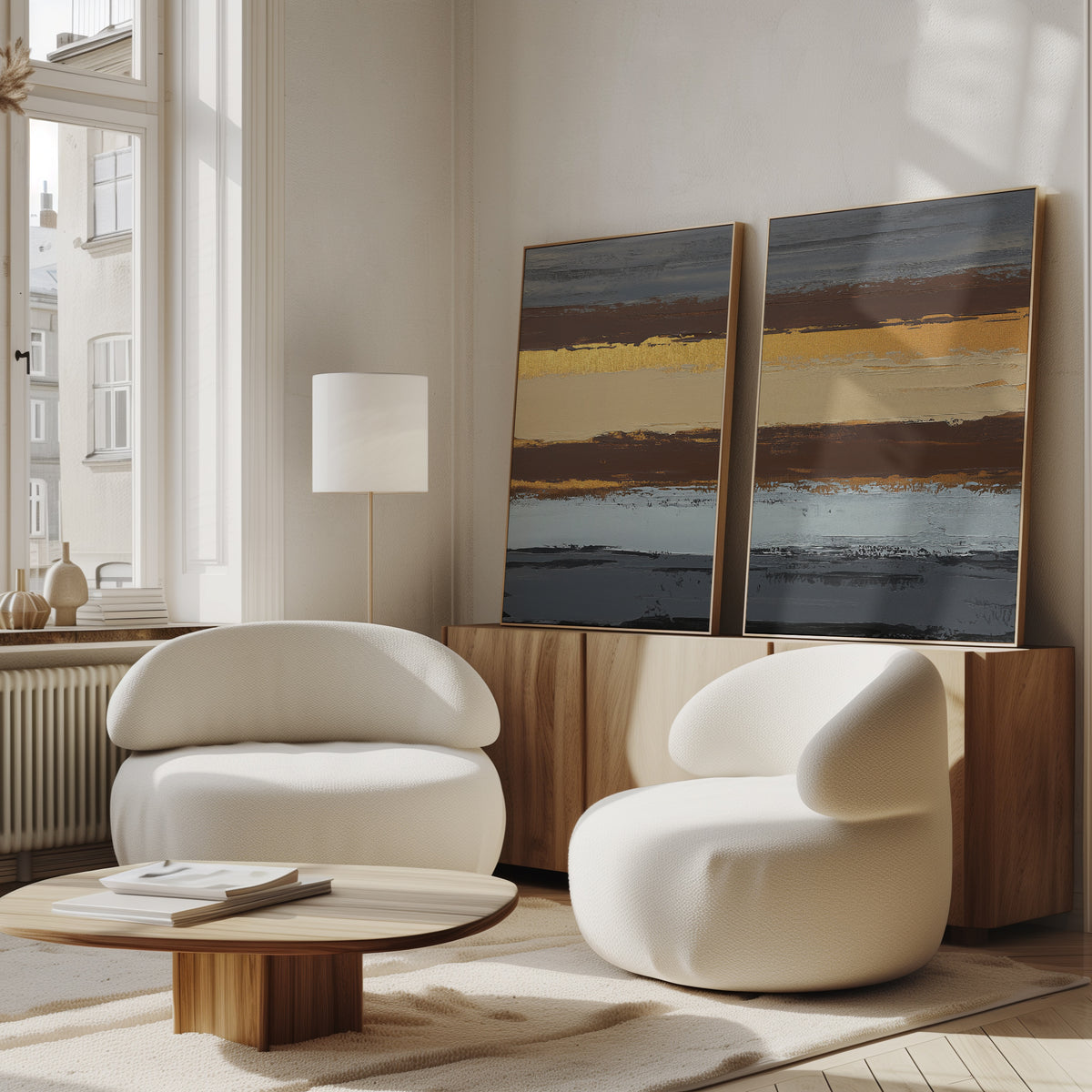 A modern living room featuring two rounded white chairs, a low wooden coffee table, and earthy vibes, with the Coastal Strata - Set Of 2 abstract prints displaying horizontal stripes on a sideboard by a large window.