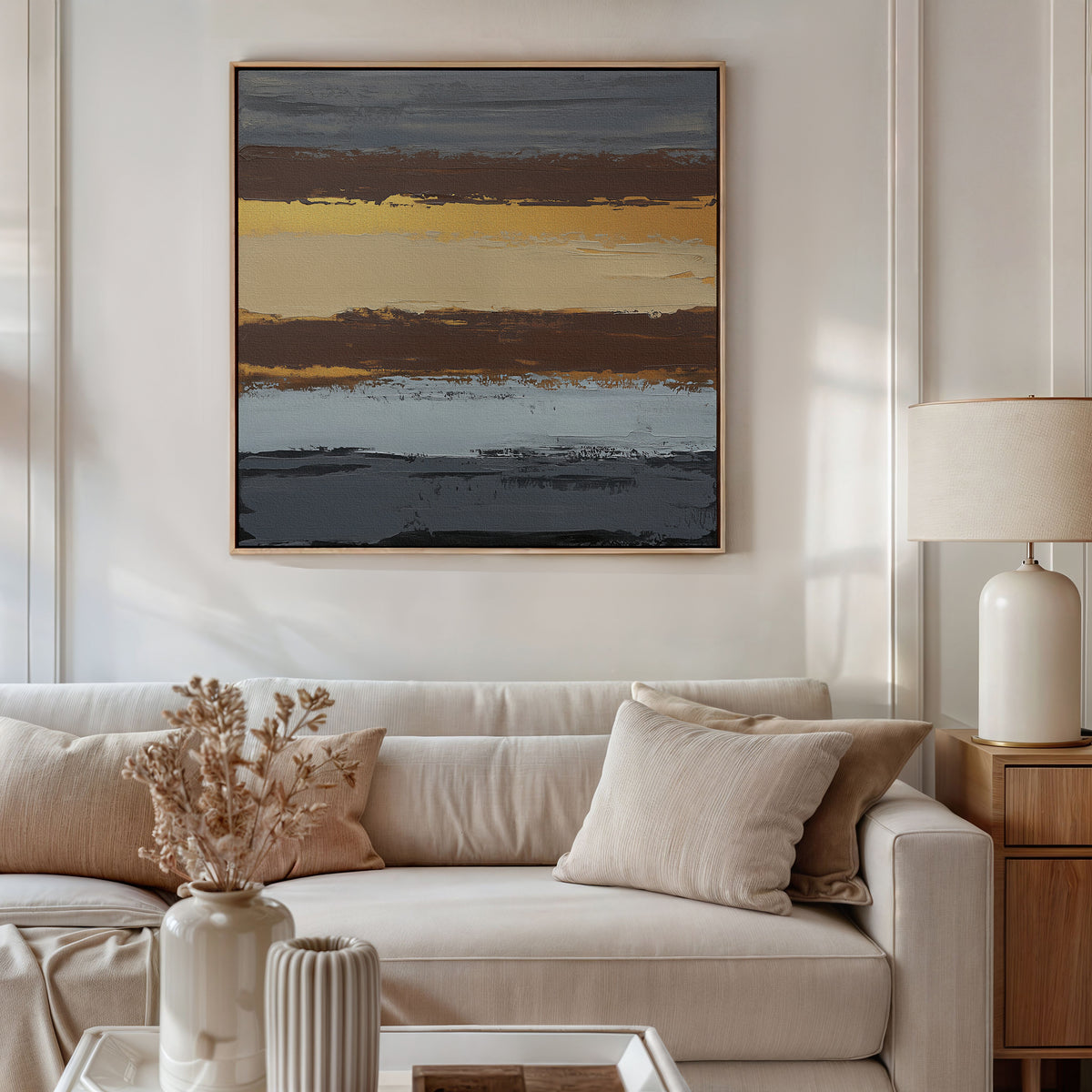 A modern living room with a beige sofa, neutral pillows, and a wooden side table with a ceramic lamp. Above the sofa hangs the Coastal Strata Canvas Art - Square, its earthy stripes in dark gray, gold, brown, and blue adding sophistication.