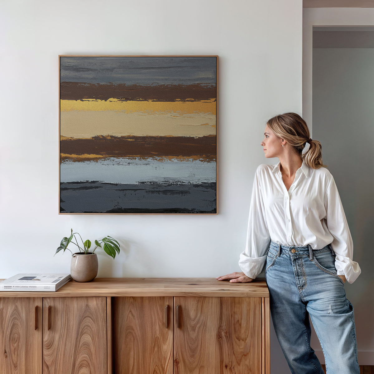 A woman in a white blouse and jeans stands by a wooden cabinet, admiring the Coastal Strata Canvas Art - Square—a contemporary piece with horizontal stripes of yellow, brown, blue, and gray—adding earthy sophistication beside a plant and books.