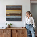 A woman in a white blouse and jeans stands by a wooden cabinet, admiring the Coastal Strata Canvas Art - Square—a contemporary piece with horizontal stripes of yellow, brown, blue, and gray—adding earthy sophistication beside a plant and books.