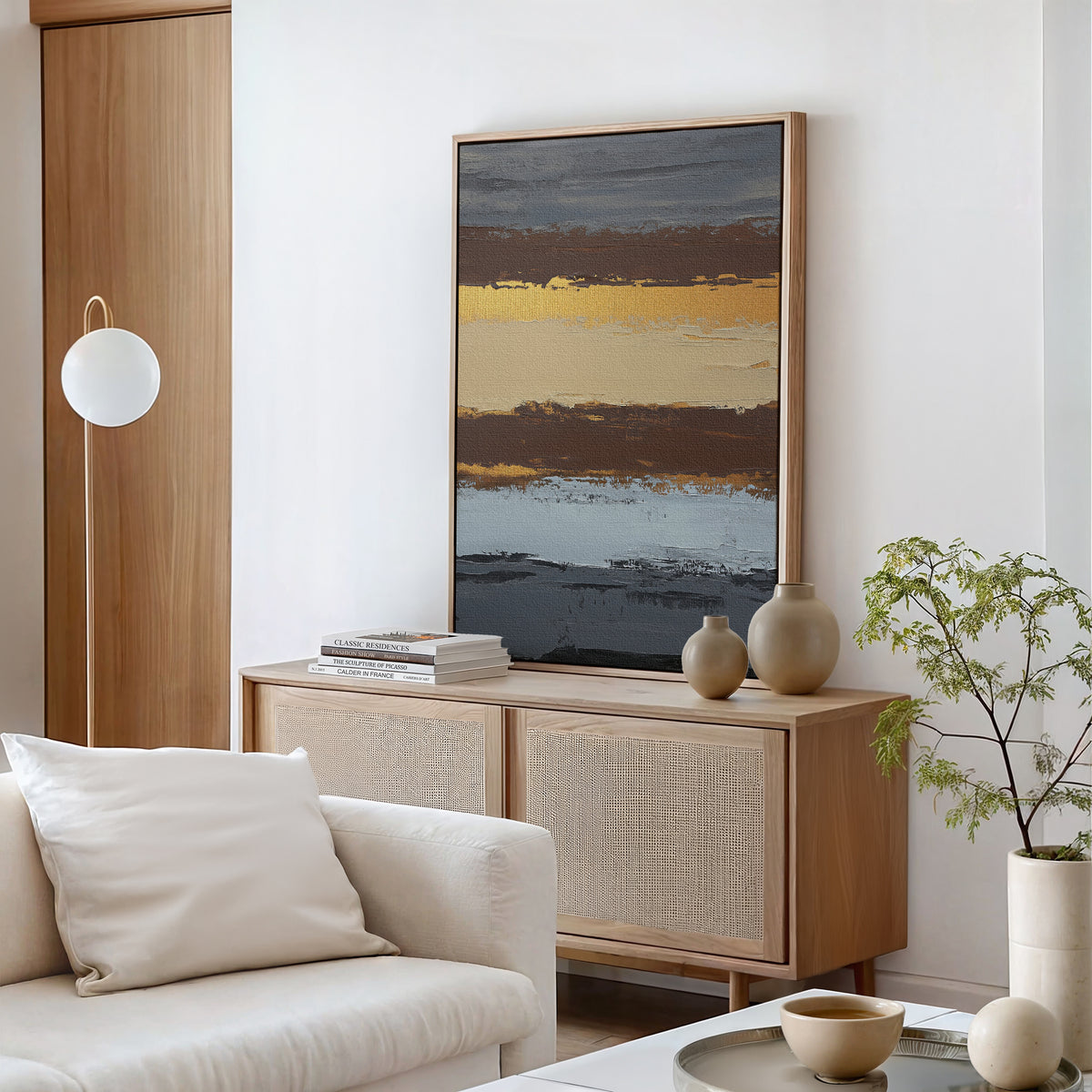 A modern living room features a light wood sideboard with neutral vases, books, and the Coastal Strata Canvas Art - Vertical, displaying gold, brown, and blue stripes for earthy sophistication beside a white sofa and potted green plant.