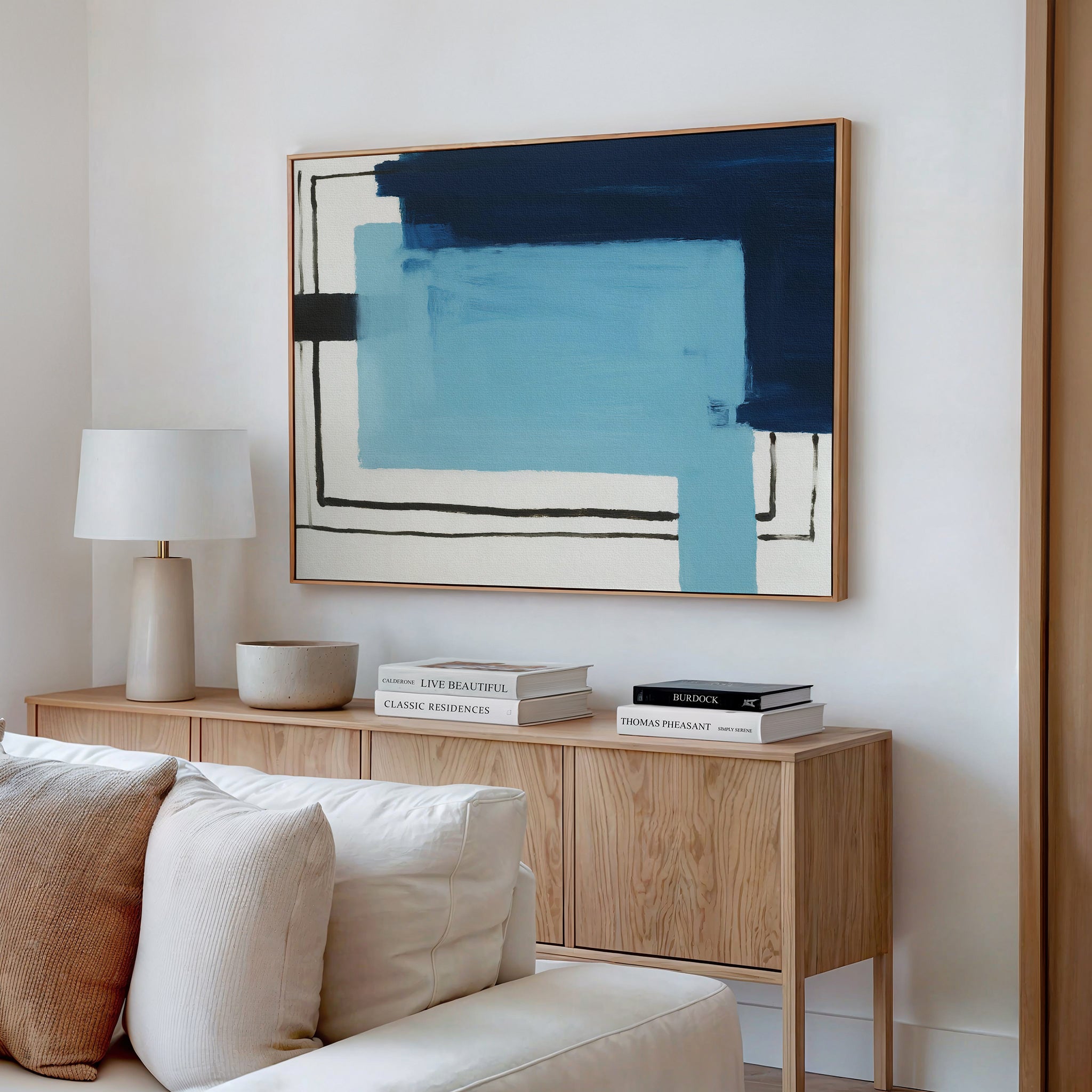 A modern living room features a wooden sideboard with books, a lamp, and a bowl. Above it hangs the Cobalt Intersection Canvas Art - Horizontal in blue and black tones. A white sofa is partly visible in the foreground.