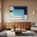 A modern living room featuring a wooden cabinet, the Cobalt Intersection Canvas Art - Horizontal in blue and black, a beige sofa, wooden coffee table with books, small plant, and soft natural light—a perfect blend of modern decor.