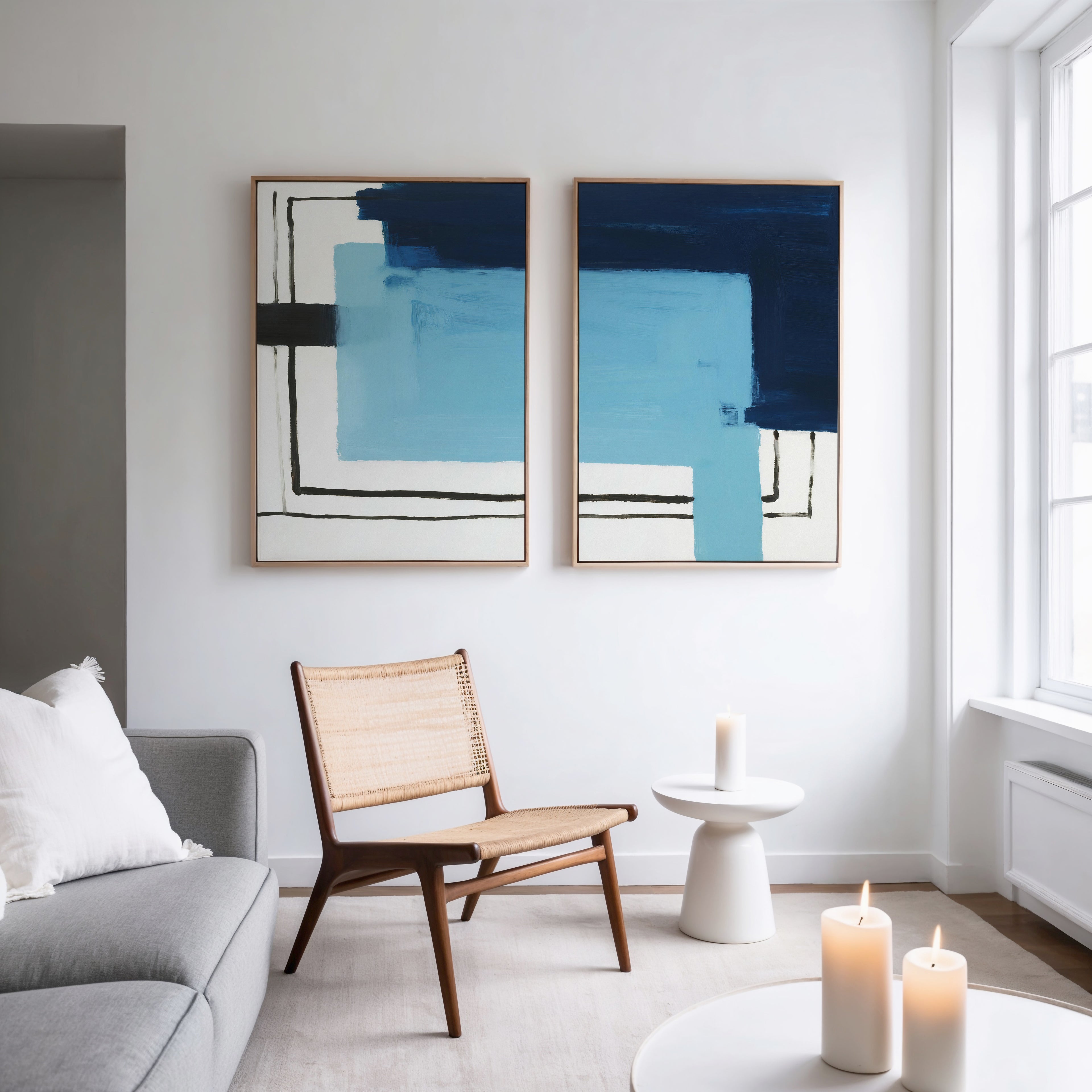 A modern living room features a wooden chair, round white side table, three lit candles, gray sofa with white pillow, and Cobalt Intersection - Set Of 2 wall art above. Natural light from large windows enhances the stylish decor.