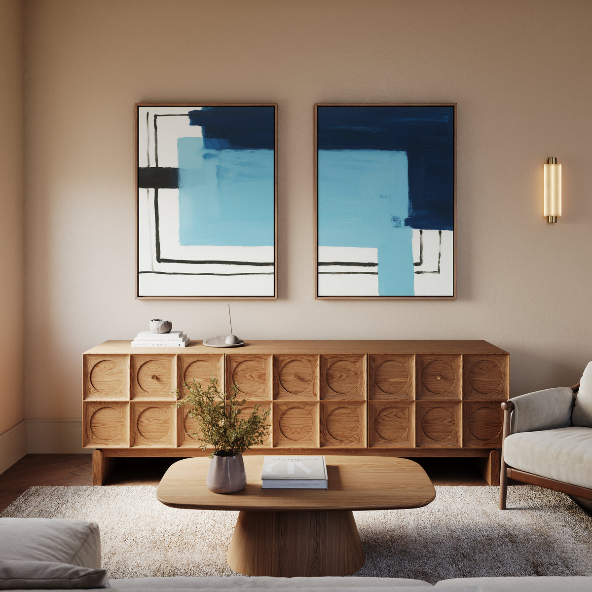 A modern living room features a wooden sideboard, a round coffee table with books and a vase, a light armchair, and the Cobalt Intersection - Set Of 2 geometric wall art above. Soft lighting enhances the cozy atmosphere.