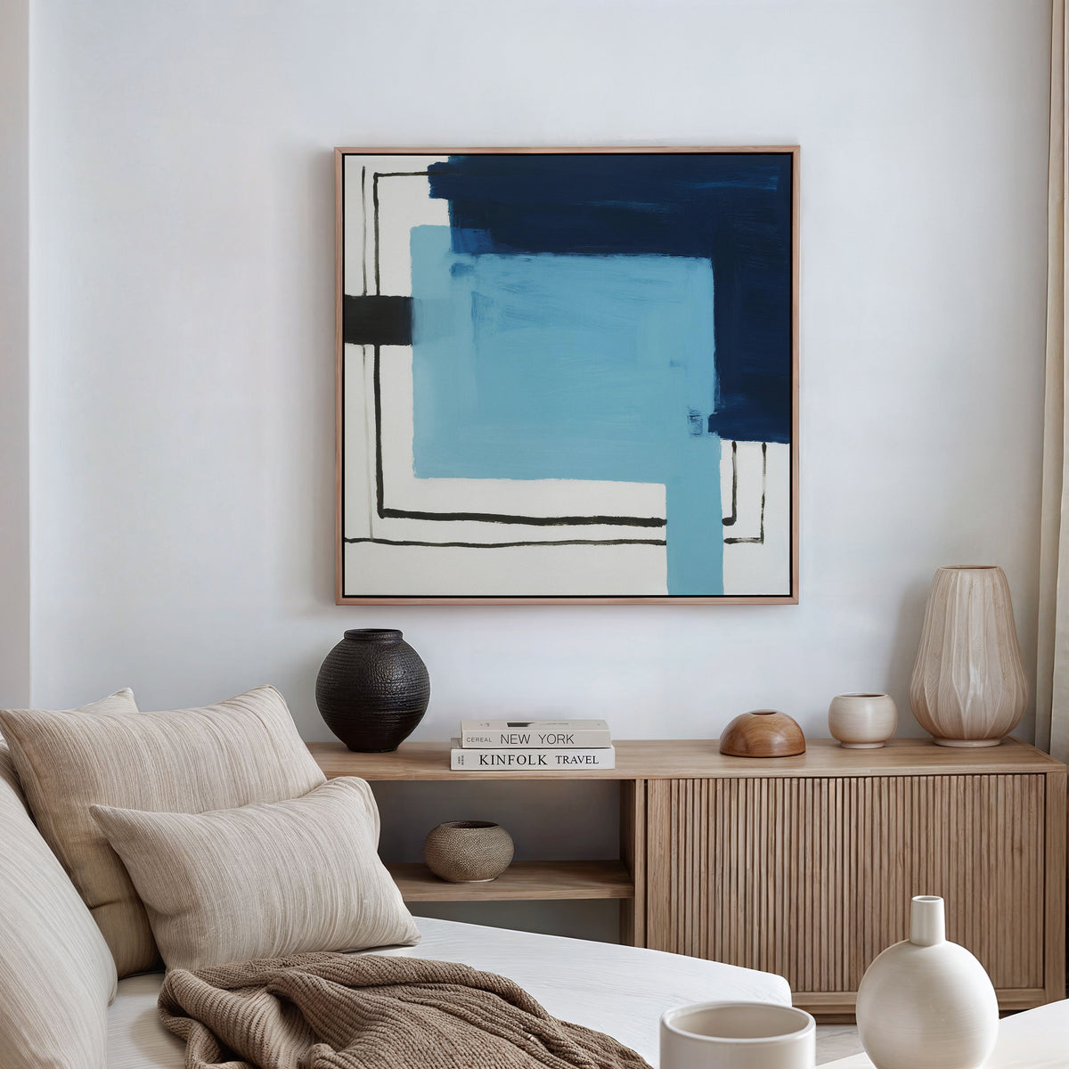 A modern living room featuring a light wood sideboard, neutral decor, and a white sofa with beige pillows. On the wall is the Cobalt Intersection Canvas Art - Square, a contemporary abstract print in blue, black, and white.