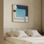 A beige bedroom features a tufted headboard, white bedding, and pillows. Above the bed hangs the Cobalt Intersection Canvas Art - Square, showcasing blue geometric shapes and black lines. A bare branch sits on a side table by the window.